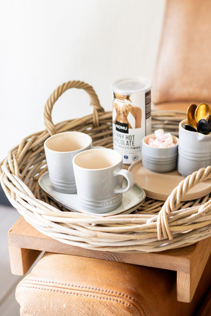 Rattan Tray with two white cups, a container of hot chocolate mix, and other small items on a wooden surface.