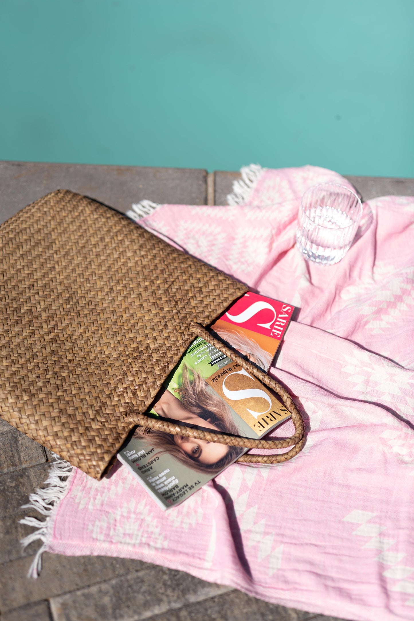 Brown woven bag with a pink towel and magazine on a poolside surface