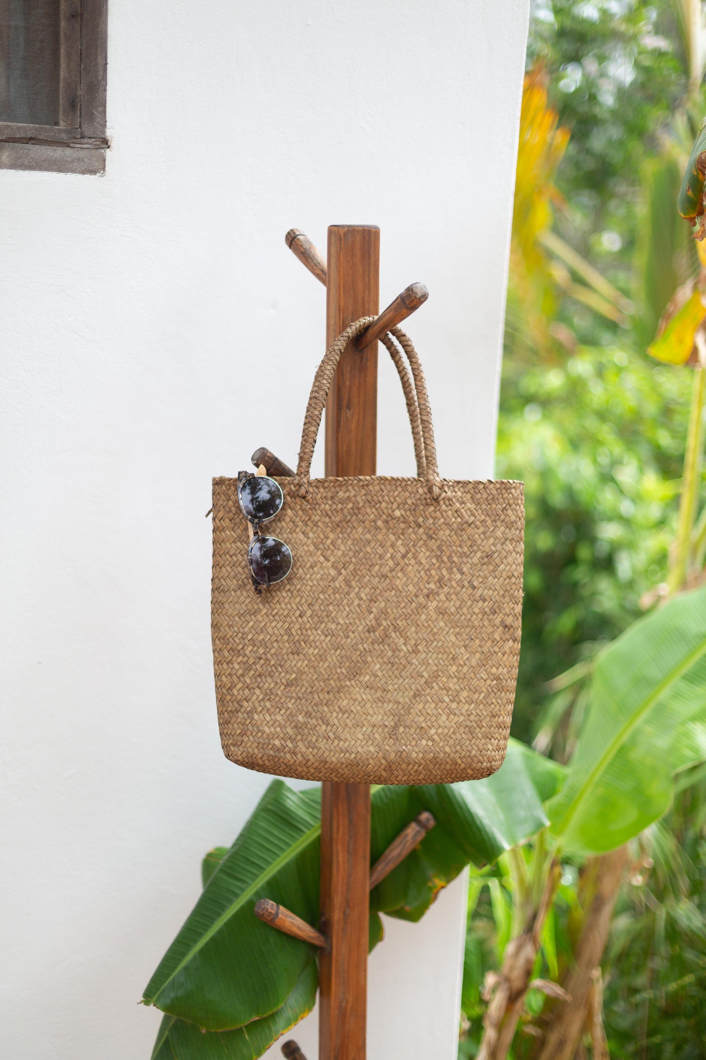 Woven bag with sunglasses on a wooden hook against a white wall with greenery.